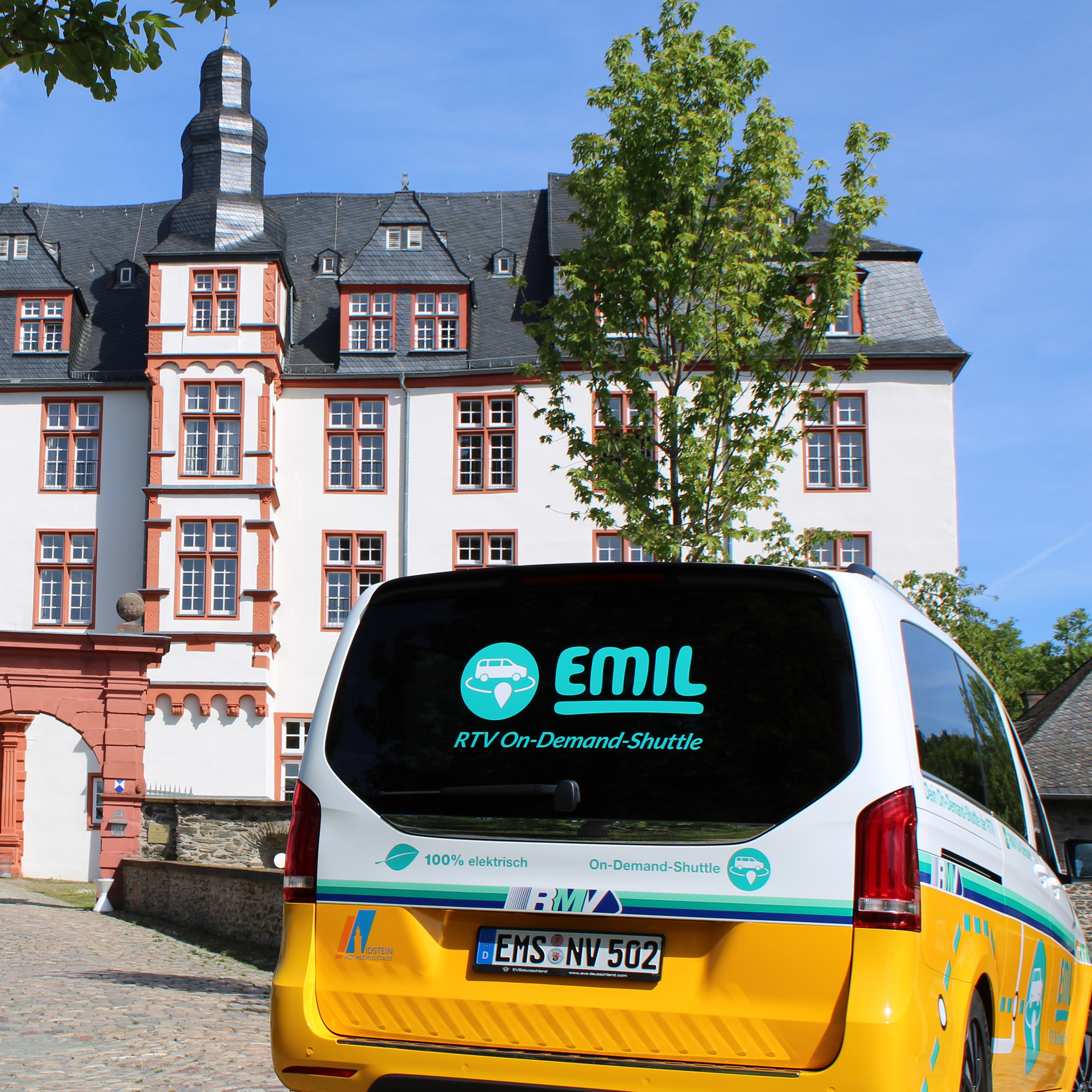 Das elektrisch betriebene On-Demand-Shuttle „EMIL“ der Rheingau-Taunus-Verkehrsgesellschaft steht vor dem historischen Idsteiner Schloss mit weiß-roter Fassade und markantem Schieferdach.