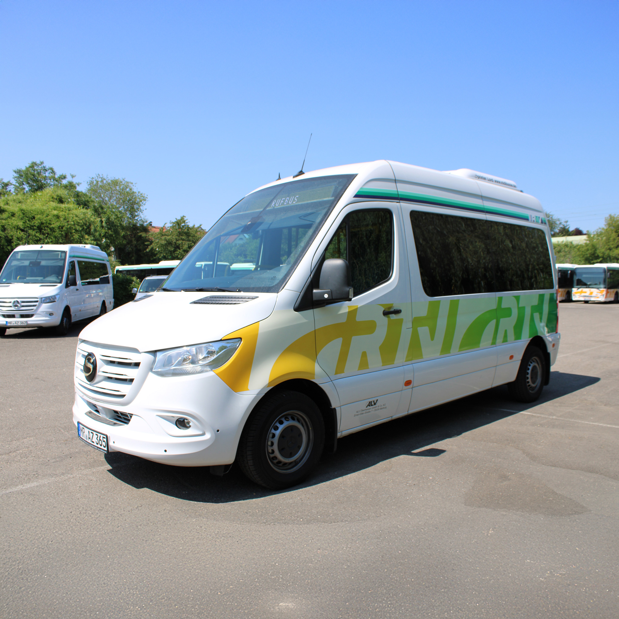 Der RTV-Rufbus für den Rheingau steht auf einem asphaltierten Platz, umgeben von Bäumen und anderen Kleinbussen. Das Fahrzeug zeigt das typische grün-gelbe Design und gehört zur Flotte des flexiblen Nahverkehrs im ländlichen Raum.
